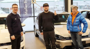 Tre personer står mellom to biler i en forhandler. Personen i midten gir tommel opp. Et «Møller Bil»-banner er synlig i bakgrunnen.