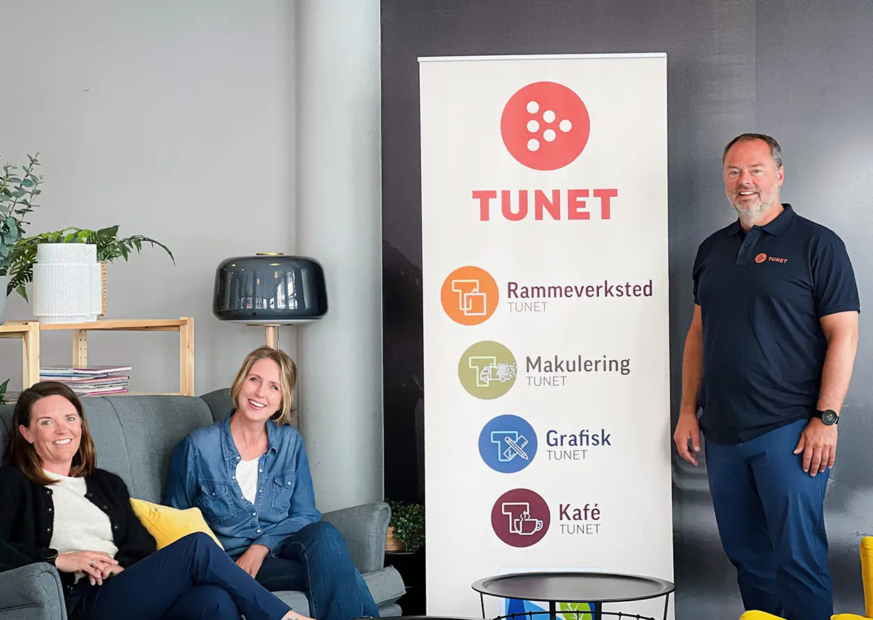 Tre personer er sett i nærheten av et banner som viser logoen og tjenestene til Tunet, inkludert rammeverksted, makulering, grafiske tjenester og en kafé. To av dem sitter på en sofa, og en står.
