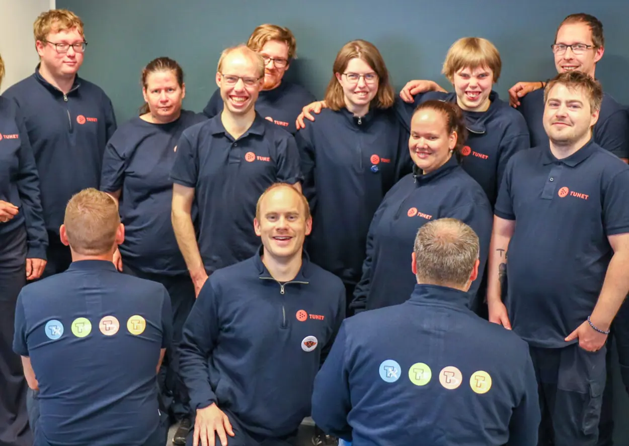 En gruppe mennesker iført mørkeblå uniformer med firmalogoer, smiler og poserer på et gruppebilde mot blå bakgrunn. Noen har ryggen vendt og viser flere logoer.