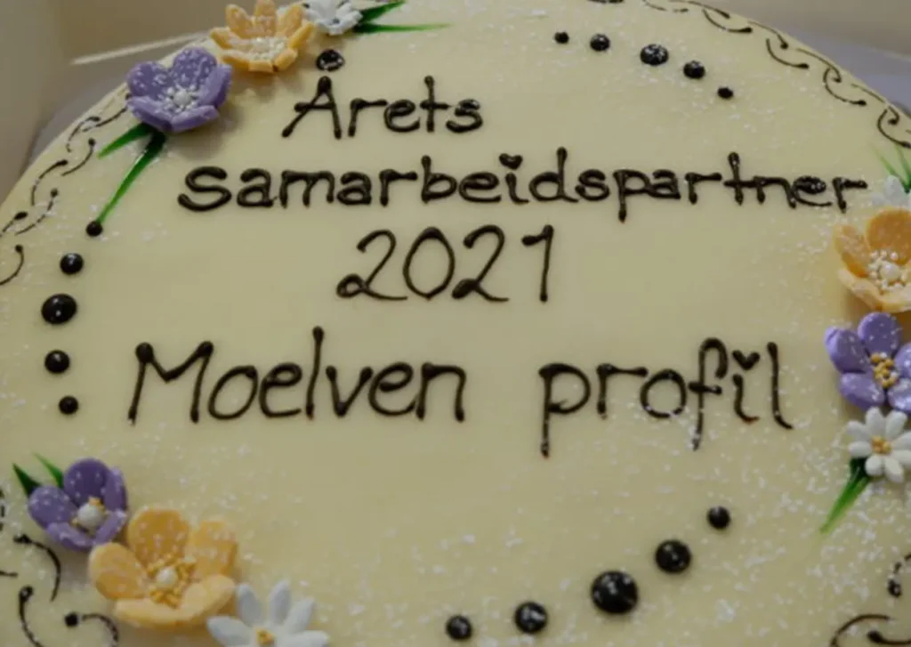 En hvit kake med glasurpynt og teksten "Årets samarbeidspartner 2021 Moelven profil" skrevet på toppen. Blomster og prikker laget av glasur pryder kantene, og feirer Tunet Elverums samarbeid med Moelven.