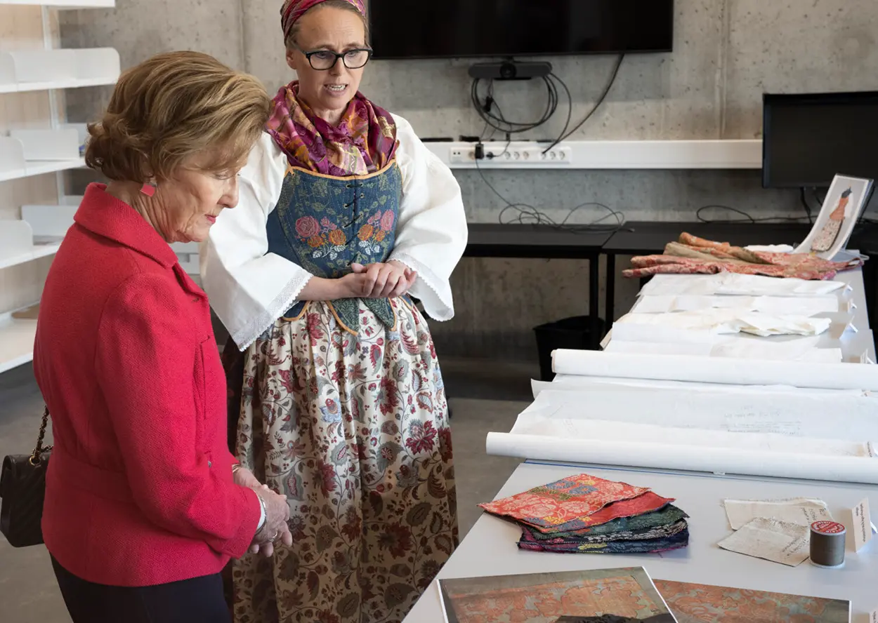 To kvinner står ved et bord med tekstiler. En kvinne, iført en tradisjonell kjole, forklarer stoffene; den andre lytter oppmerksomt, iført rød jakke. Ulike stoffer og mønstre er spredt på bordet, som gjenspeiler det nitidige håndverket til Agena Tunet i Elverum tilrettelagt arbeid, beundret selv av Dronning Sonja.