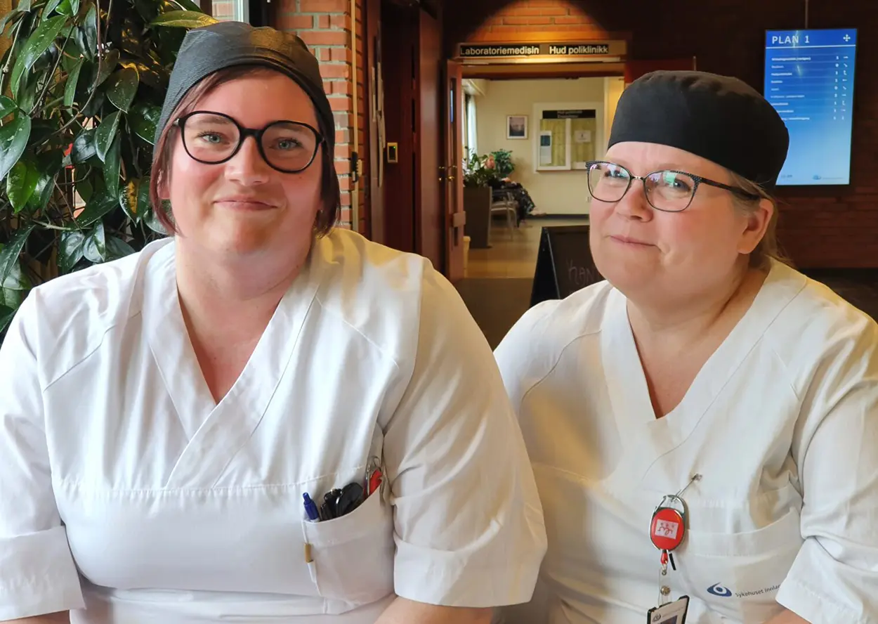 To personer iført hvite uniformer og svarte capser sitter side om side i en gang på Tunet Elverum og smiler. Begge har briller og penner i brystlommene. Et skilt i bakgrunnen angir laboratorie- og radiologiområdet, og viser kompetansebrevet de tjente for tilrettelagt arbeid.