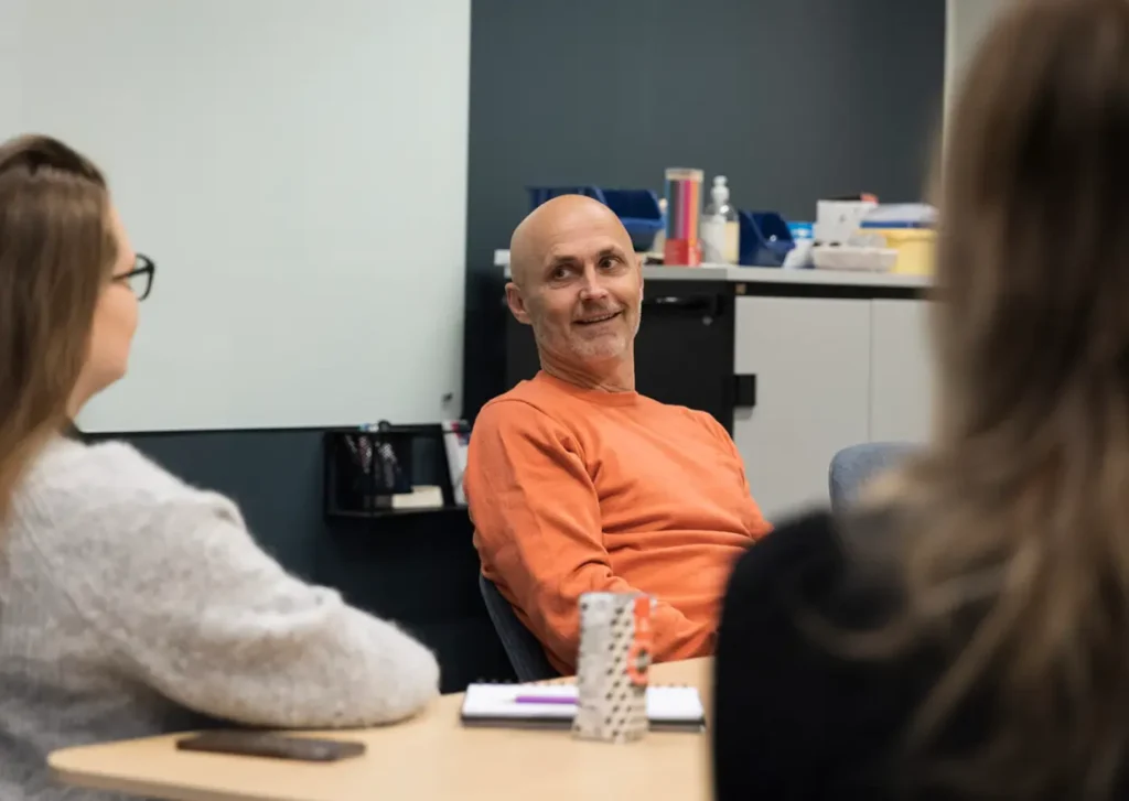 En skallet mann i oransje skjorte sitter ved et bord og diskuterer karriereveiledning med to kvinner. Det er en tavle og noen gjenstander på en hylle i bakgrunnen.