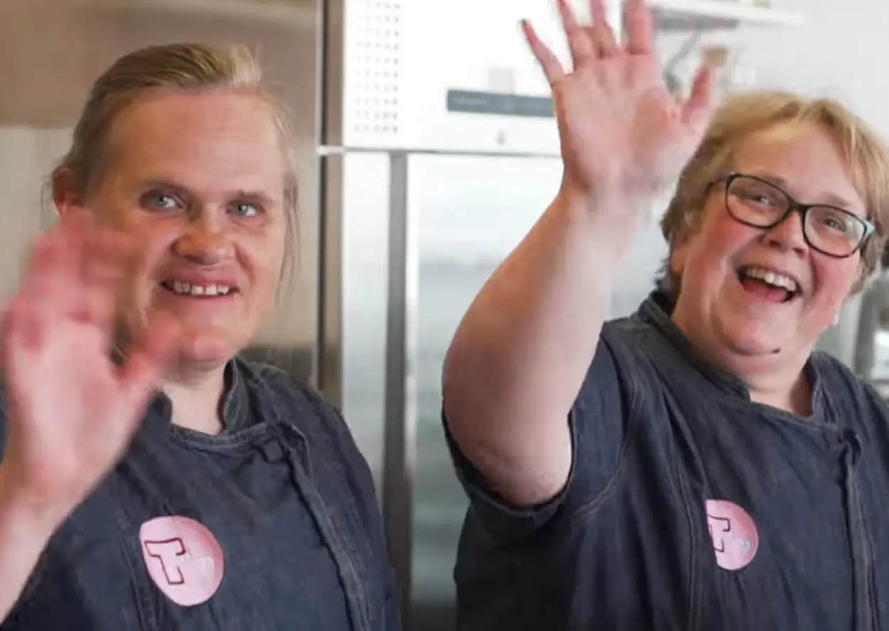 To personer iført mørke skjorter med rosa merker smiler og vinker til kameraet i en kjøkkensetting, og ser ut som de er bak kulissene på en musikkvideo-shoot.