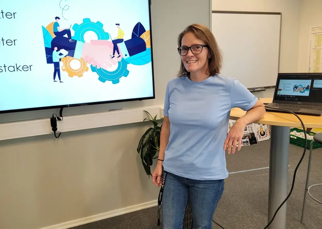 En kvinne står og smiler ved siden av en presentasjonsskjerm og en bærbar datamaskin på et bord. Skjermen viser en håndtrykkgrafikk med tannhjul og tegneseriepersoner. Som opplæringskoordinator er hun uformelt kledd i lyseblå t-skjorte og jeans.