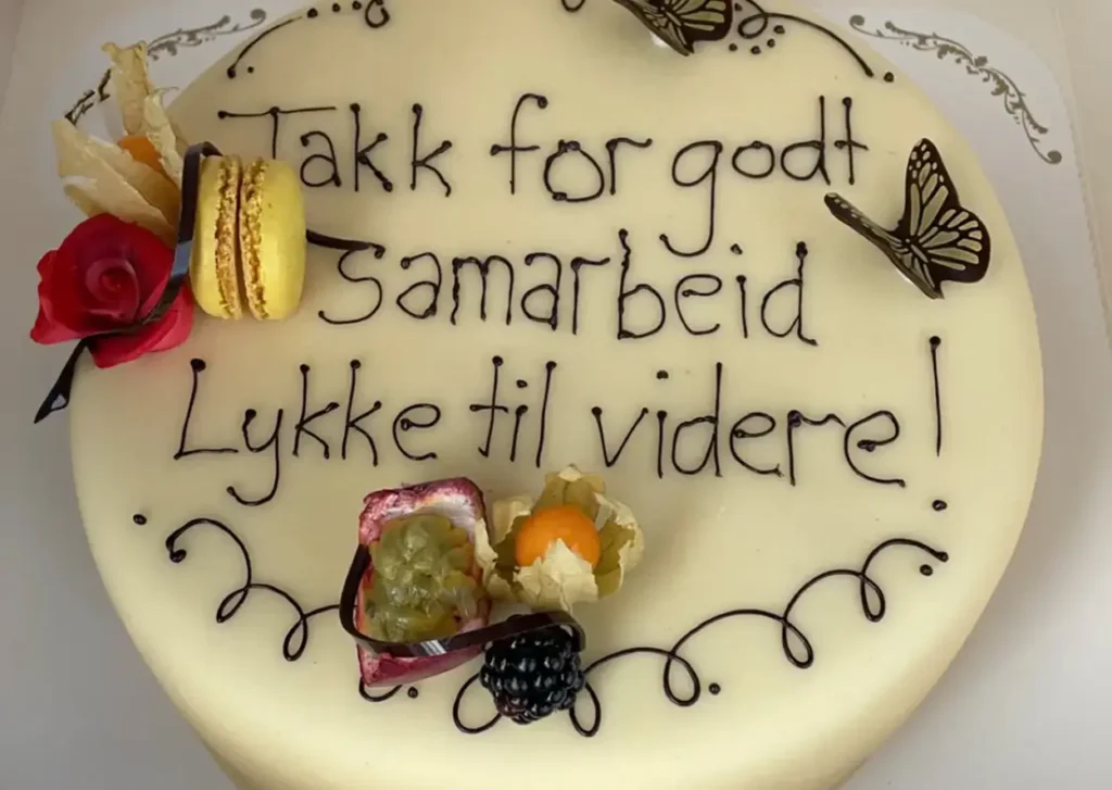 En kake med hvit glasur, dekorert med blomster, en makron, frukt og sommerfuglfigurer. Kaken har norsk tekst skrevet i sjokolade, takker for godt samarbeid og ønsker gry lykke til i ny jobb.