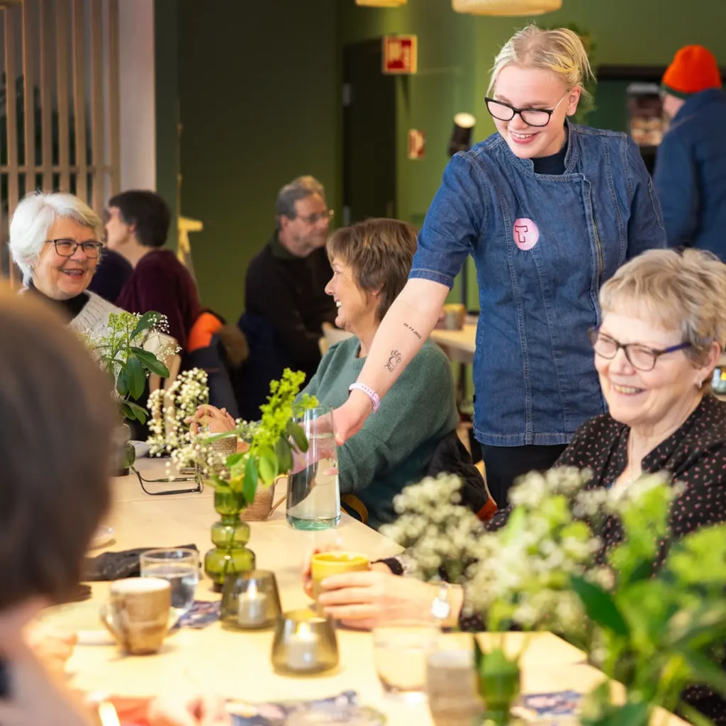 En smilende server i dongeriforkle skjenker vann til en gruppe sittende eldre kvinner ved et bord pyntet med grønne vaser, hvite blomster og krus på Tunet Kafe. Folk sosialiserer seg i bakgrunnen i uformelle omgivelser.