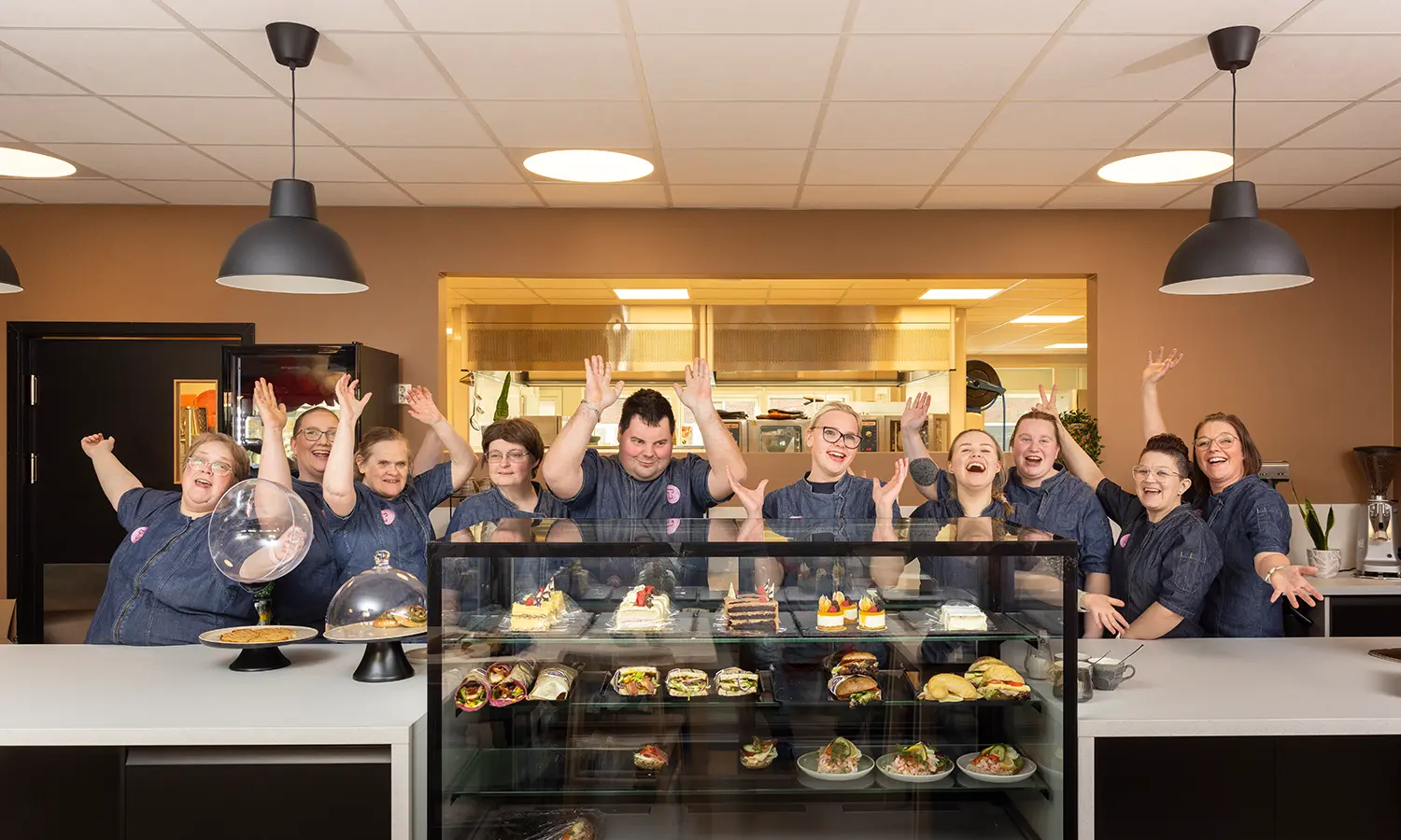 En gruppe mennesker fra Tunet Elverum står bak en glassmonter fylt med ulike matvarer, smiler og rekker opp hendene i en feiringsgest. De har på seg matchende blå uniformer.