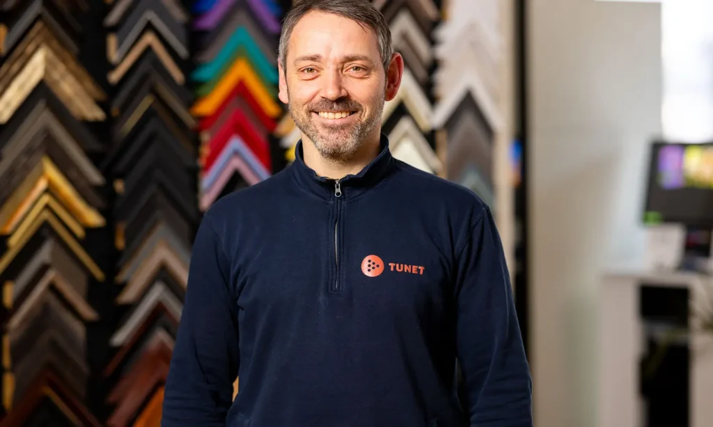 En mann iført en mørkeblå skjorte med logoen "TUNET" står foran en fargerik utstilling av assorterte rammehjørner. Han smiler og vender mot kameraet, og gir fra seg en følelse av ekspertise som om han skulle diskutere makulering.
