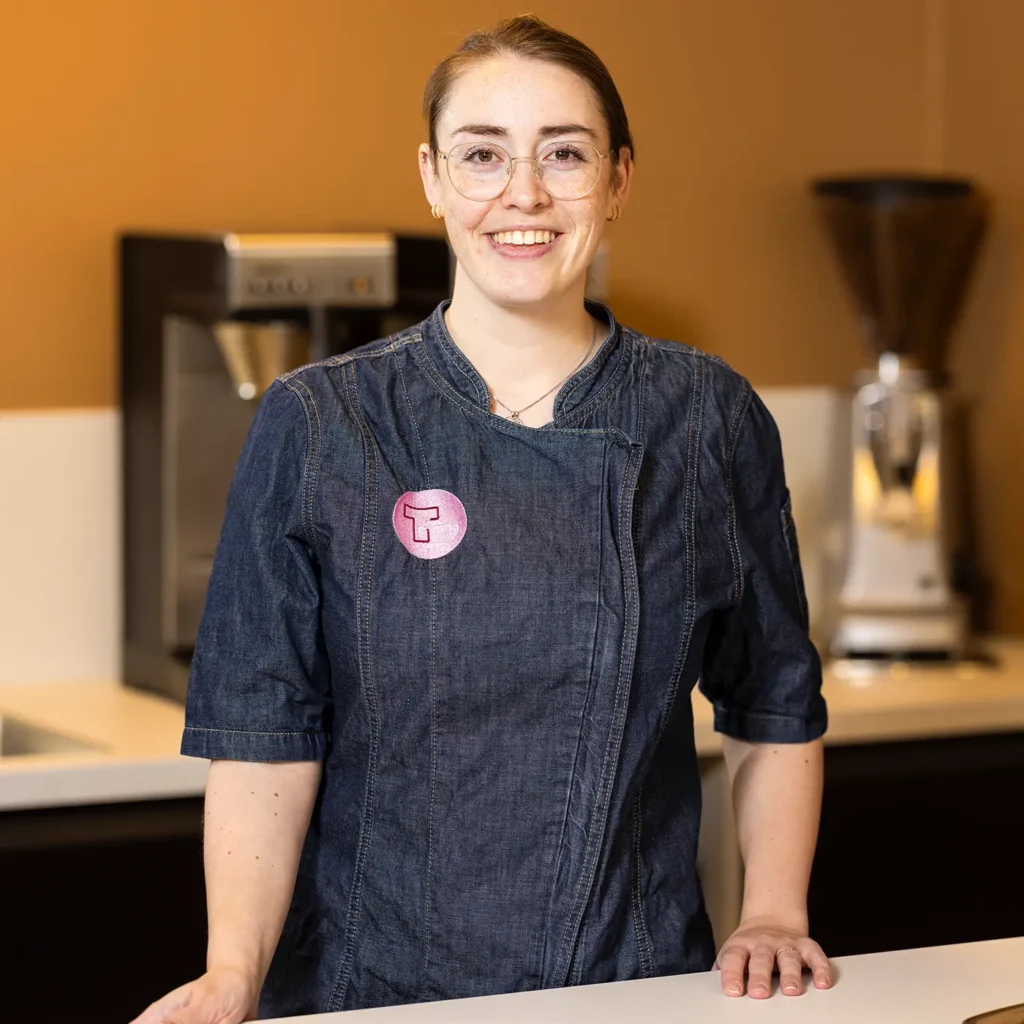 En person med briller og denim kokkefrakk med rosa logo står og smiler på et kjøkken med kaffekvern og annet utstyr. For henvendelser, kontakt oss.