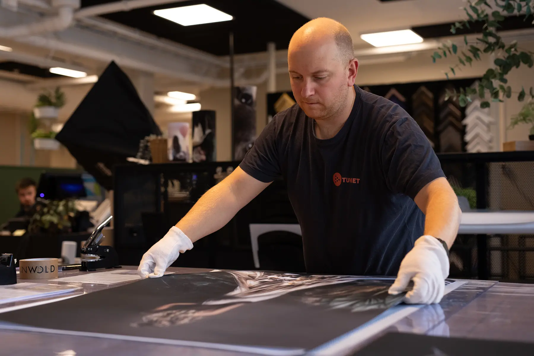 En mann med hansker håndterer forsiktig et stort fotografi på et bord i et moderne arbeidsområde, og forbereder det for trykk og ekspedisjon av bestillinger i nettbutikken til den anerkjente kunstneren Linn Wold.