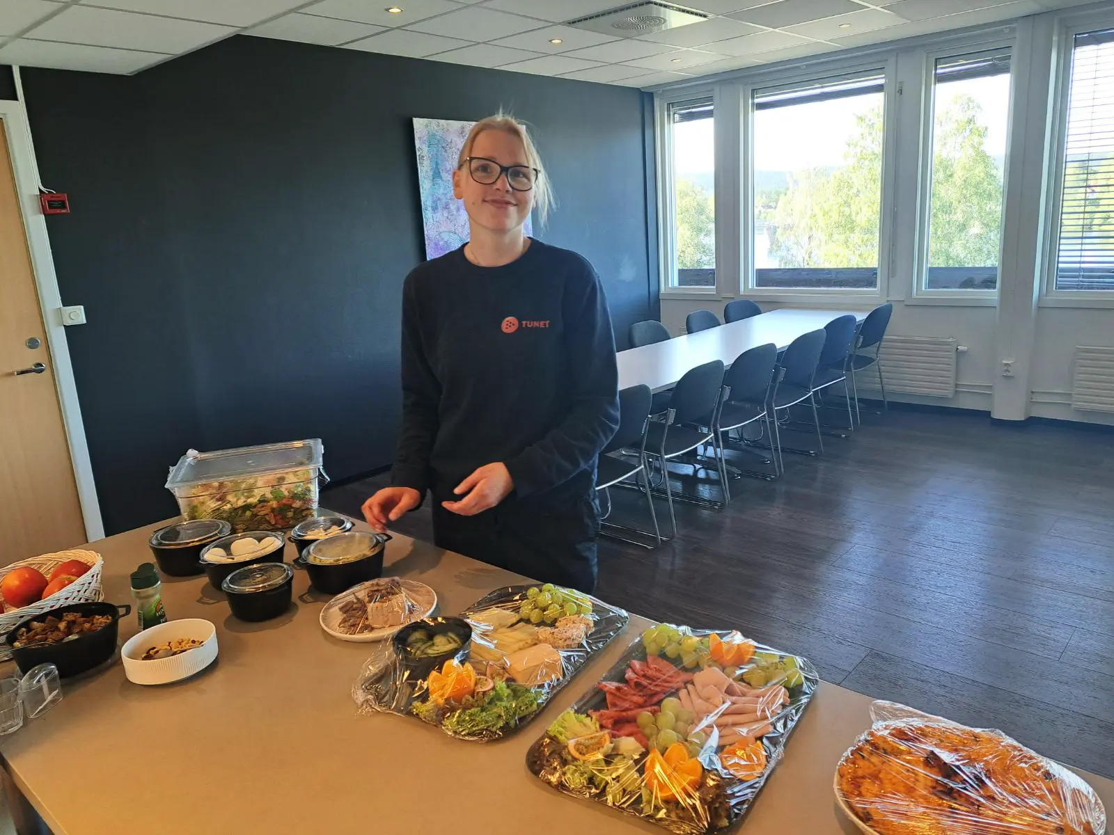 En person står ved et bord i konferanserommet TUNET KAFÈ – MIDT I LEIRET, omgitt av diverse fat med frukt, ost og kjøtt.